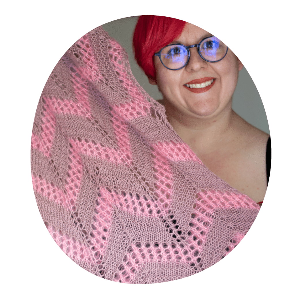 Eine Frau mit roten Haaren und lilaner runder Brille hält lächelnd ein Strickstück in die Kamera, das ein Zackenmotiv in rosa und altrosa hat.