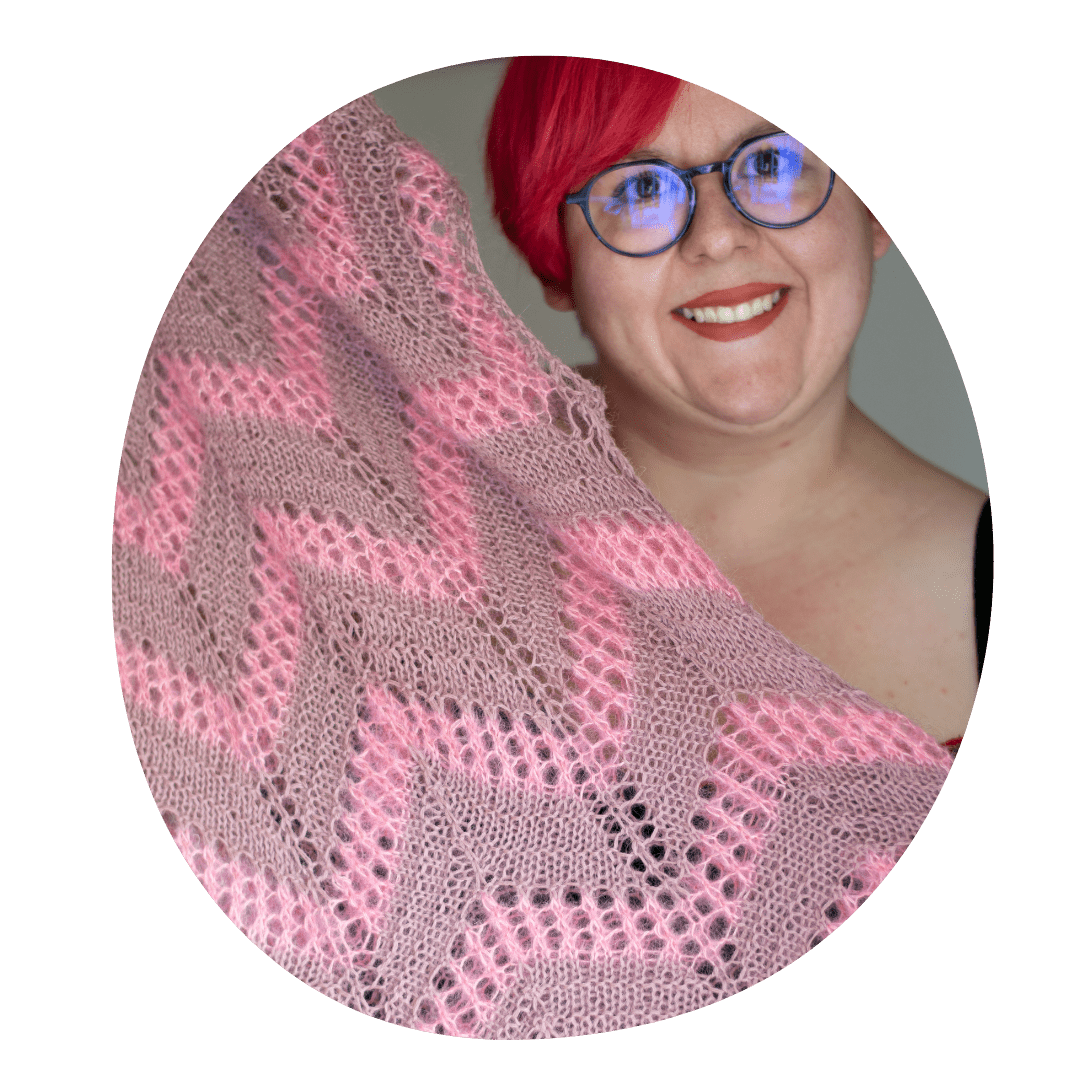 Eine Frau mit roten Haaren und lilaner runder Brille hält lächelnd ein Strickstück in die Kamera, das ein Zackenmotiv in rosa und altrosa hat.