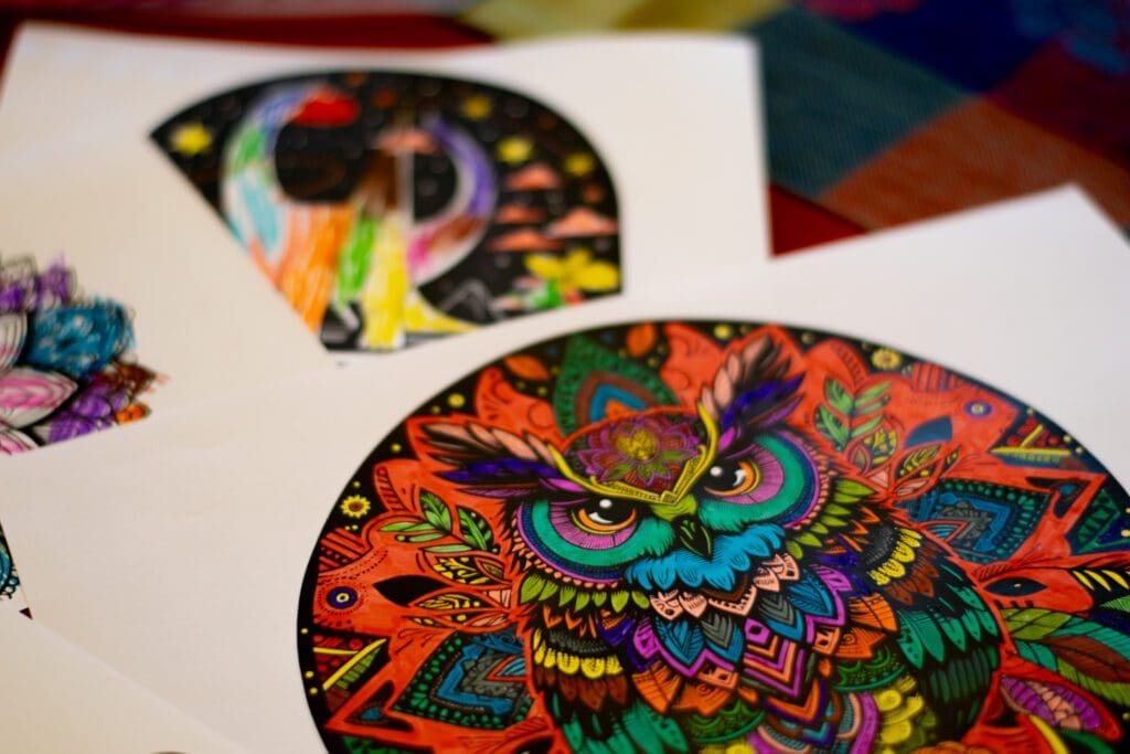 3 bunte Ausmalbilder auf weißem Papier, Im Hintergrund unscharf ein Dino-Motiv und ein Blumen-Mandala, im Vordergrund scharf zu sehen ein Eulenmandala.