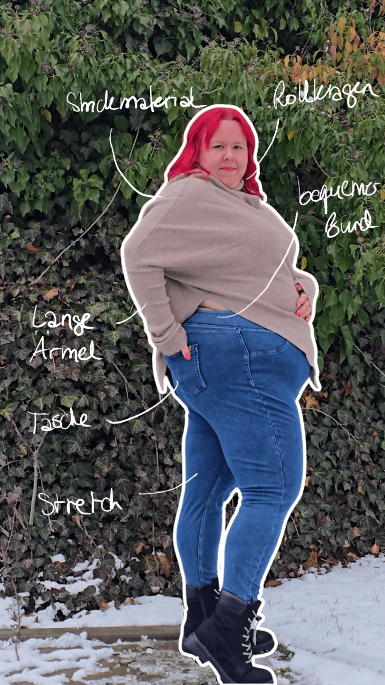 Dicke rothaarige Frau trägt schwarze Stiefel, eine mittelblaue Jeans und einen beigen Poncho Pullover. Sie steht seitlich und hält die Hand in der hinteren Hosentasche.