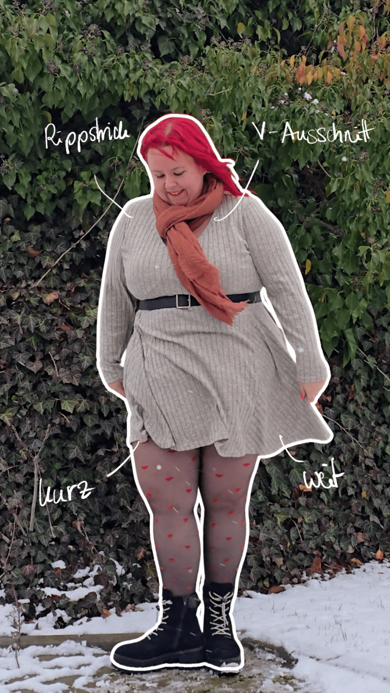 Dicke rothaarige Frau trägt schwarze Stiefel, eine Strumphose mit roten Herzen, ein Strickkleid in beige, einen schwarzen Gürtel und einen rotbraunen Schal.