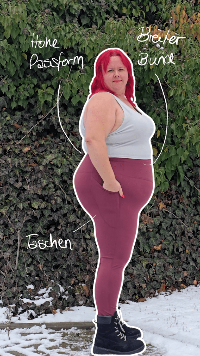 Dicke rothaarige Frau trägt schwarze Stiefel, eine weinrote Hose und ein weißes Top. Sie steht seitlich und hält die sichtbare Hand in der Hosentasche.